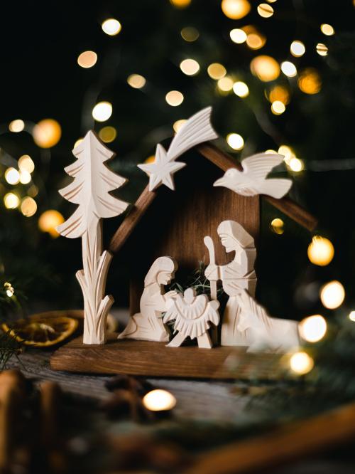 Drewniana szopka na tle choinki z zapalonymi lampkami.