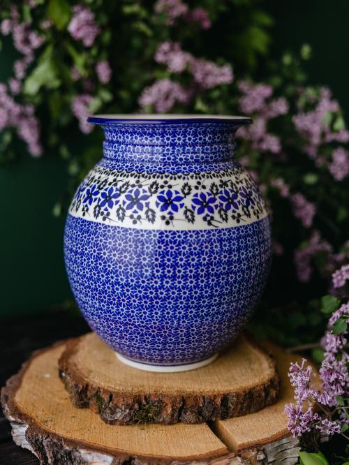 Wielki, ceramiczny wazon uchwycony przodem stojąc na drewnianych deseczkach na tle fioletowych bzów.