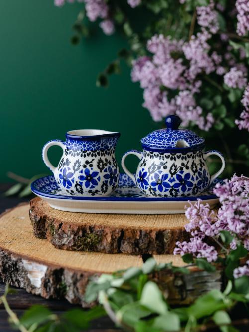 Ceramiczny komplet w postaci mlecznika i cukiernicy umieszczony na drewnianej deseczce wśród fioletowych bzów.