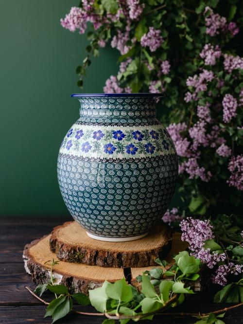 Wielki, ceramiczny wazon uchwycony przodem stojąc na drewnianych deseczkach na tle fioletowych bzów.