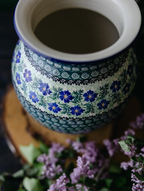 Zbliżenie na ceramiczny wazon uchwycony delikatnie z góry stojąc na drewnianych deseczkach na tle fioletowych bzów.