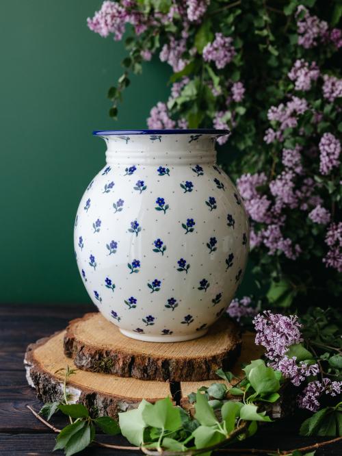 Wielki, ceramiczny wazon uchwycony przodem stojąc na drewnianych deseczkach na tle fioletowych bzów.