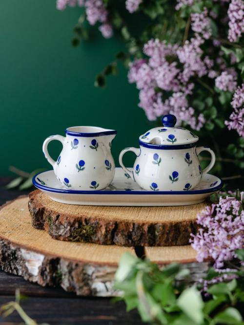 Ceramiczny komplet w postaci mlecznika i cukiernicy umieszczony na drewnianej deseczce wśród fioletowych bzów.