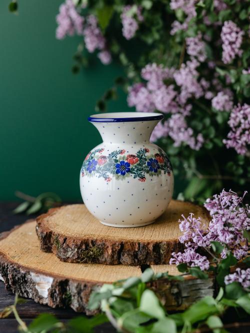 Okrągły, ceramiczny wazon uchwycony przodem stojąc na drewnianych deseczkach na tle fioletowych bzów.