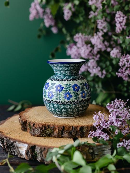 Okrągły, ceramiczny wazon uchwycony przodem stojąc na drewnianych deseczkach na tle fioletowych bzów.