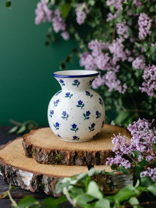 Okrągły, ceramiczny wazon uchwycony przodem stojąc na drewnianych deseczkach na tle fioletowych bzów.