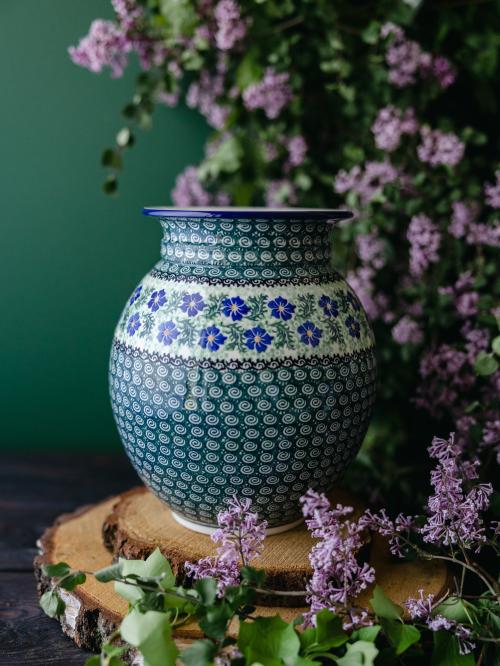 Wielki, ceramiczny wazon uchwycony przodem stojąc na drewnianych deseczkach na tle fioletowych bzów.
