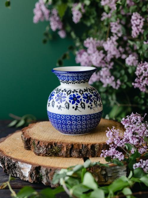 Okrągły, ceramiczny wazon uchwycony przodem stojąc na drewnianych deseczkach na tle fioletowych bzów.