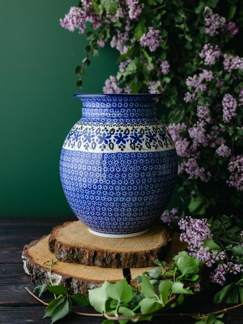 Wielki, ceramiczny wazon uchwycony przodem stojąc na drewnianych deseczkach na tle fioletowych bzów.