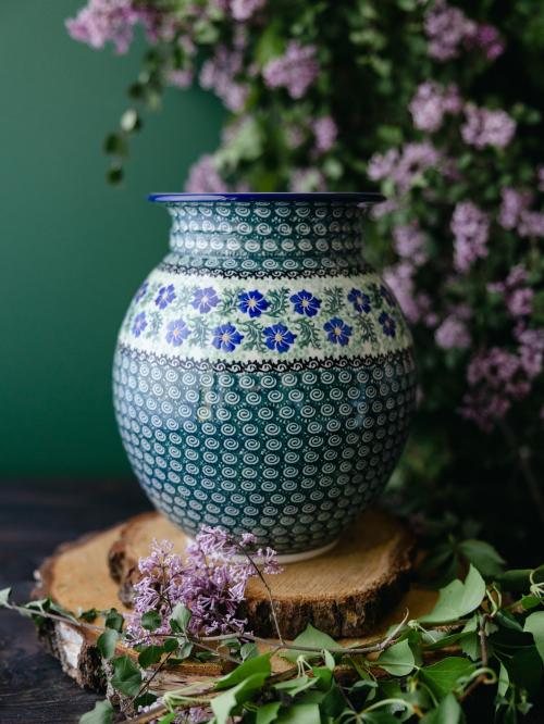 Wielki, ceramiczny wazon uchwycony przodem stojąc na drewnianych deseczkach na tle fioletowych bzów.