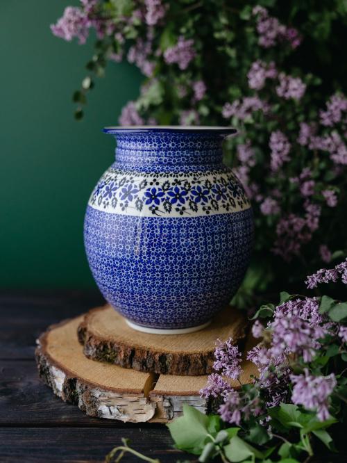 Wielki, ceramiczny wazon uchwycony przodem stojąc na drewnianych deseczkach na tle fioletowych bzów.